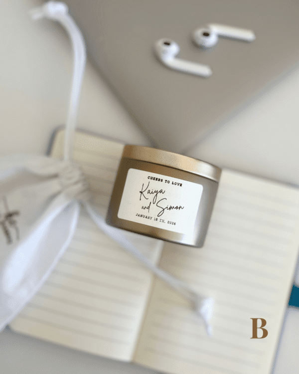 Cheers to Love personalised wedding candle bonbonniere in champagne tin with custom couple names label, resting on white cotton drawstring bag, Perth WA Krisha Essence