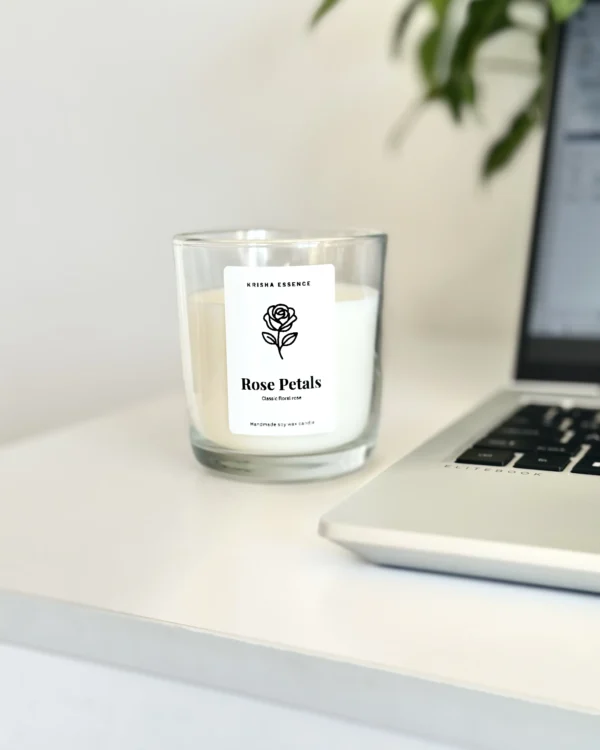 Krisha Essence Rose Petals soy jar candle sitting on a white desk next to a laptop, featuring a classic fresh cut roses candle fragrance.